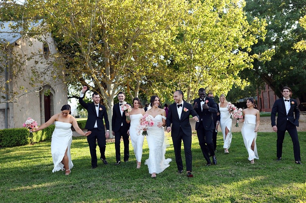 Wedding party walking on grass with trees in the background, women in white dresses and men in tuxedos, some holding bouquets.