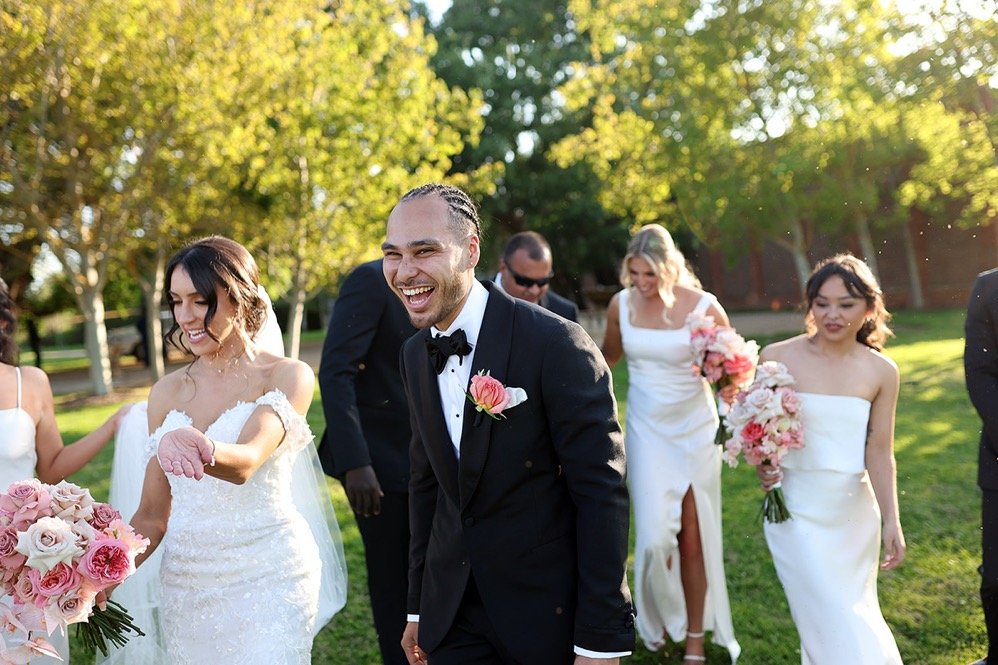 A group of wedding guests, including a bride in white gown and a groom in black tuxedo, celebrate outdoors on a sunny day with trees in the background.