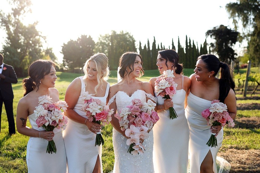 Five women in white dresses holding bouquets, standing outdoors on a sunny day, smiling and talking to each other, with trees and a grassy area in the background.