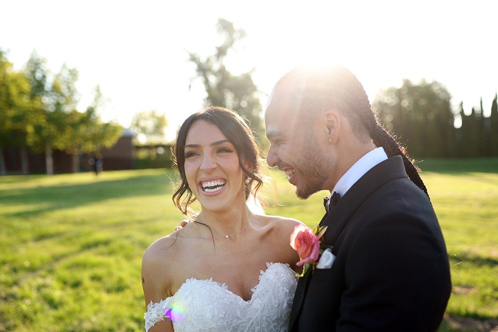 Bride and groom on their wedding day outdoors, smiling and laughing with sunlight behind them.
