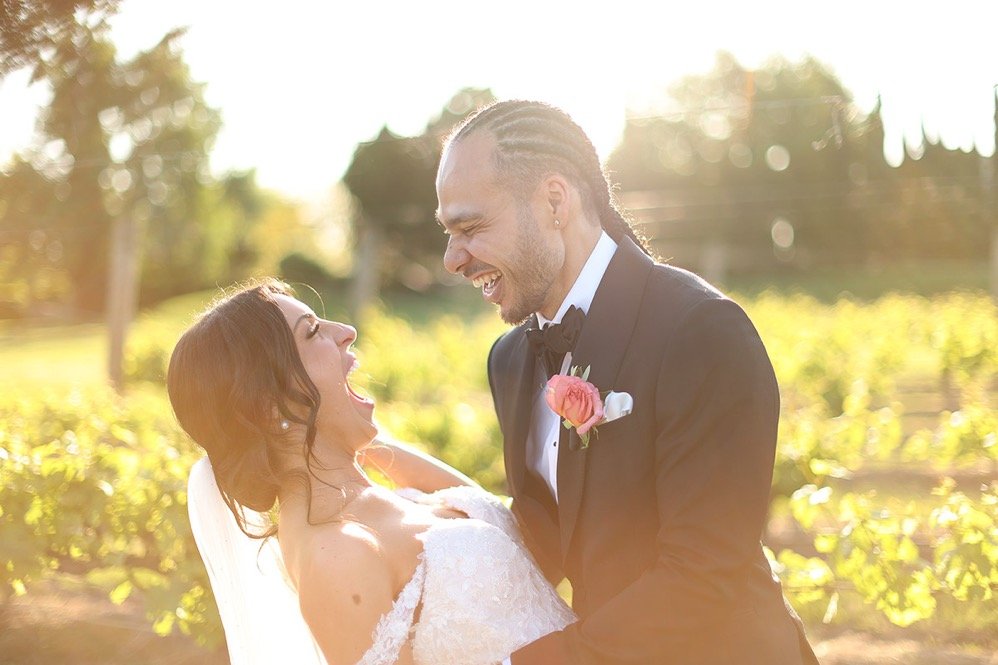 A happy couple on their wedding day, outdoors in a sunny garden or vineyard with greenery in the background.