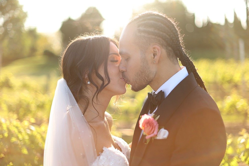 A bride and groom sharing a kiss outdoors during sunset, with the bride wearing a white wedding dress and veil, and the groom in a tuxedo with a pink boutonniere.