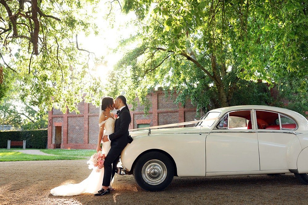 A bride and groom stand close together in front of a vintage white car under a large, leafy tree. The bride wears a white wedding dress and holds a bouquet, while the groom is dressed in a black suit. They appear to be about to kiss or share an intim