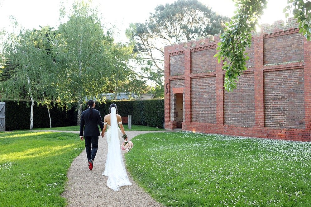 A bride and groom walking hand in hand on a curved gravel path in a lush green garden, with a red brick building and trees in the background.