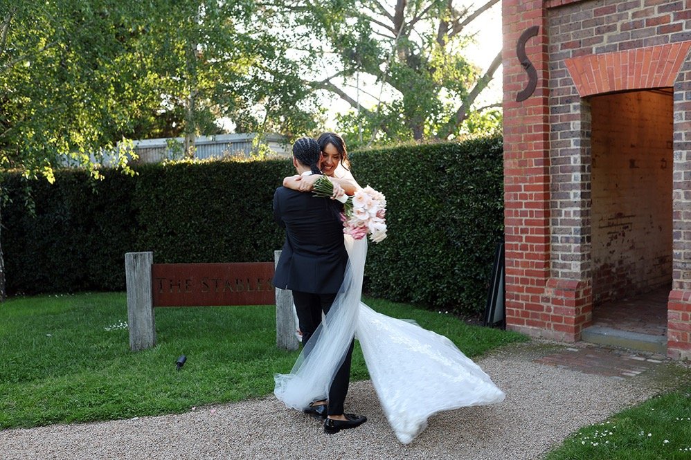 Bride in a wedding dress hugging a man in a suit outdoors near a brick building and greenery.