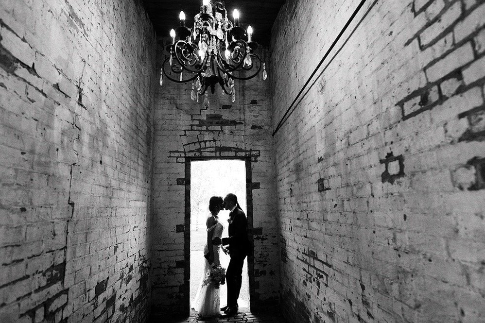 Silhouetted bride and groom standing close together in front of a bright window, inside a narrow brick-walled corridor with a chandelier hanging overhead.