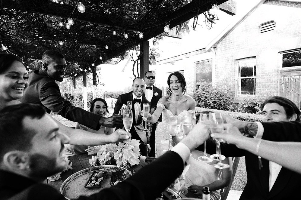 A group of people at a wedding celebration, raising glasses for a toast outdoors with string lights overhead and a brick house in the background.