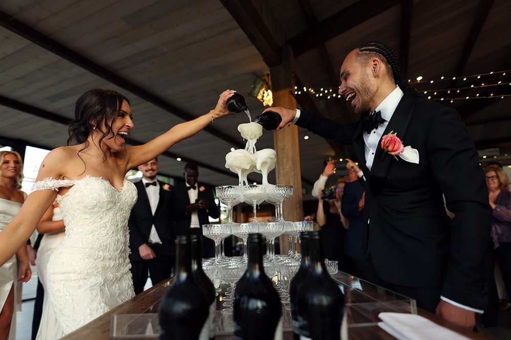 A bride and groom pour champagne into a tower of glasses at their wedding reception, with wedding guests watching in the background.