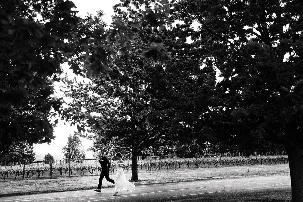 A black and white photo of a bride and groom walking hand in hand on a path in a park, surrounded by large trees.