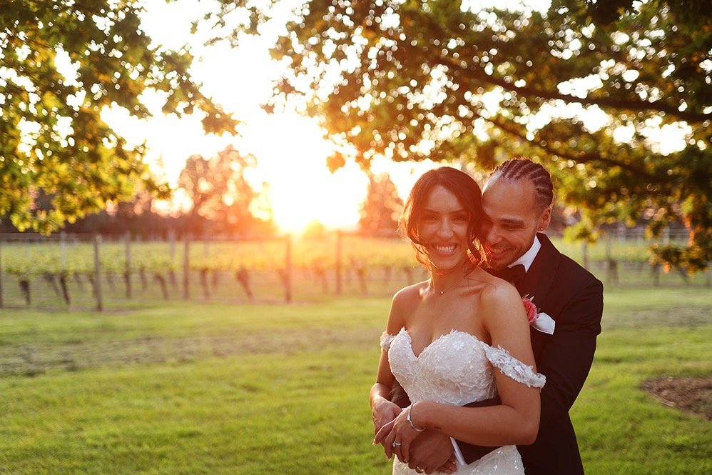 A newlywed couple smiling and embracing outdoors at sunset, with green grass, trees, and a fence in the background.