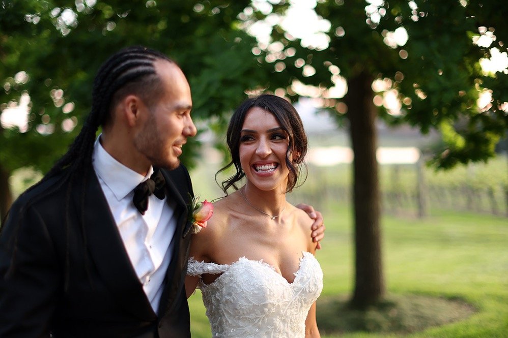 Bride and groom smiling outdoors, with the groom in a black tuxedo and the bride in a white lace wedding dress, surrounded by greenery.