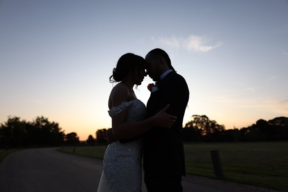 Silhouette of a couple in wedding attire embracing at sunset outside, with a sunset sky and trees in the background.
