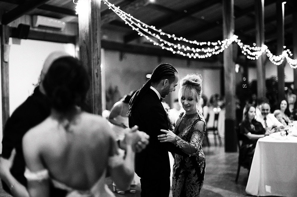 A black and white photo of a man and woman dancing closely at a social event, with others seated at a table in the background. Decorated with string lights hanging from the ceiling.