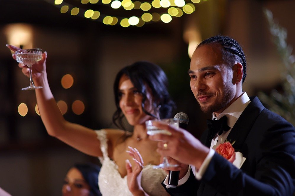 A groom in a tuxedo and a bride in a wedding dress giving a toast at their wedding reception, with a blurred background of lights.