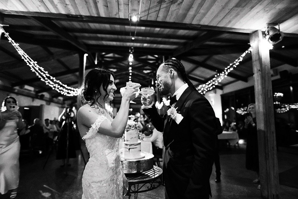 A bride and groom celebrate at their wedding reception, toasting with drinks and smiling at each other, with a wedding cake on a table between them, under string lights in a decorated indoor venue.