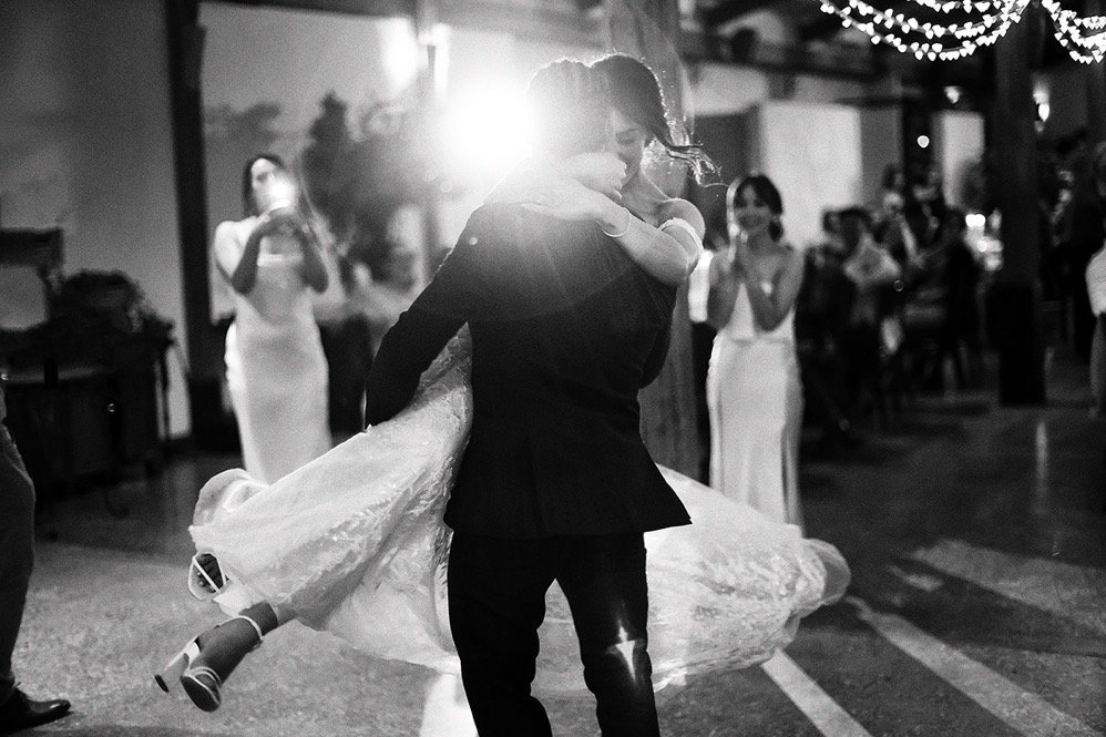 A woman in a wedding dress being lifted and danced with by a man in a tuxedo at her wedding reception, with guests watching in the background