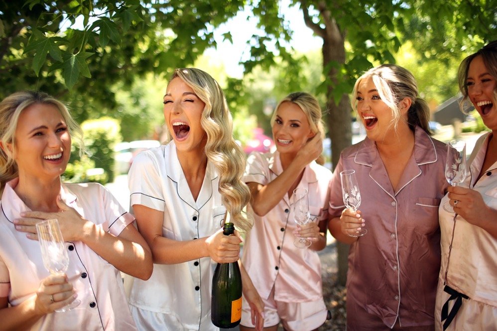 Group of women in pajamas celebrating outdoors under green trees, holding champagne glasses, smiling and laughing.