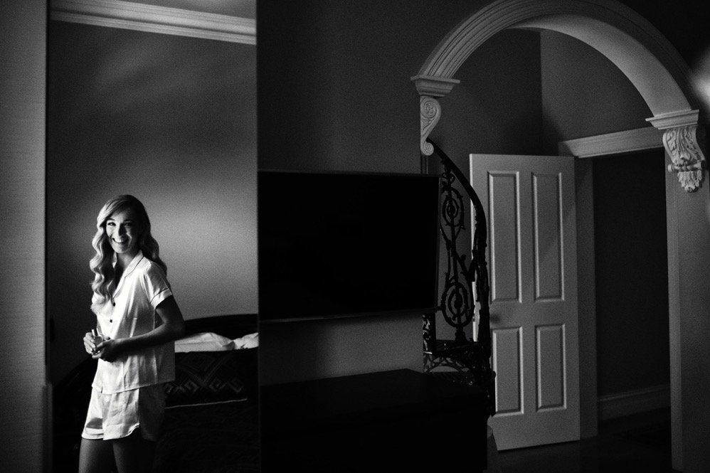 A woman with long wavy hair smiling in a bedroom, wearing light-colored pajamas, standing next to a door and a television, with an open door revealing a hallway.