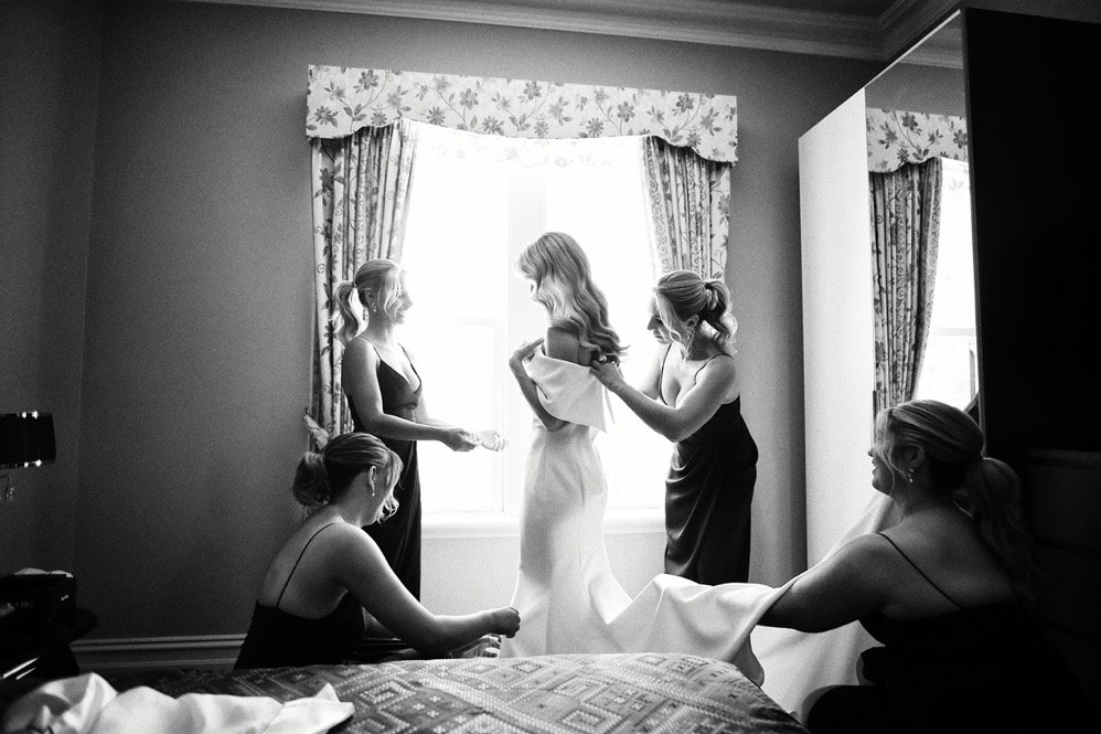 A bride in a wedding dress is getting ready with her bridesmaids in a bedroom, two of them are helping her with her dress while the other two are watching.