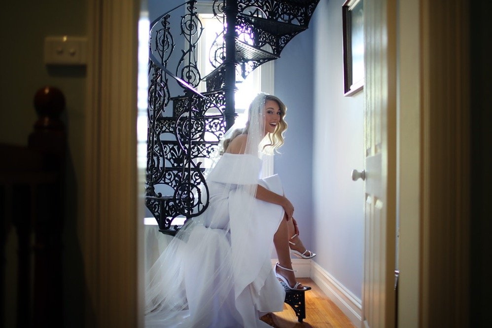Bride in a white wedding dress and veil sitting on a staircase, smiling, as she adjusts her shoe.