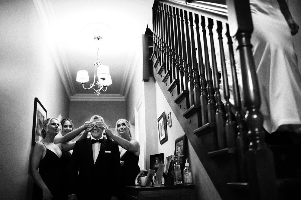 Three women in black dresses covering a man's eyes in a hallway with framed photos and a staircase.