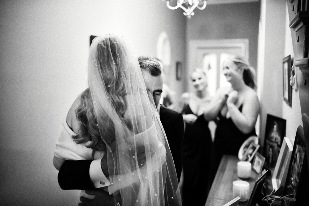 A bride and groom embrace closely in a room, with blurred family members in the background, during a wedding celebration.