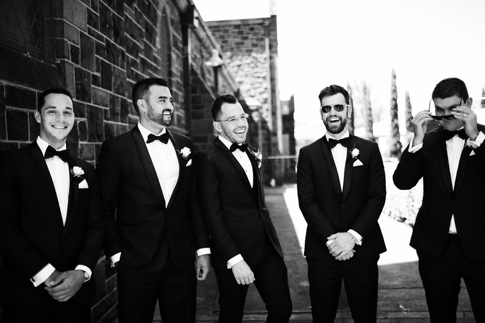 A group of five men dressed in tuxedos, standing outdoors against a brick wall, enjoying a moment of laughter and camaraderie.