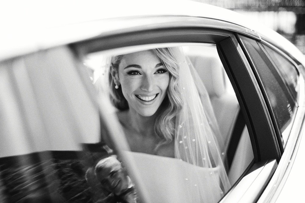 Brunette bride smiling inside a car, wearing a veil and pearl earrings, in black and white.
