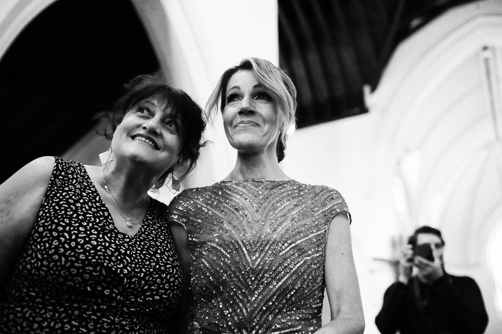 Two women smile and pose for a photo at an indoor event, with a photographer capturing the moment in the background.