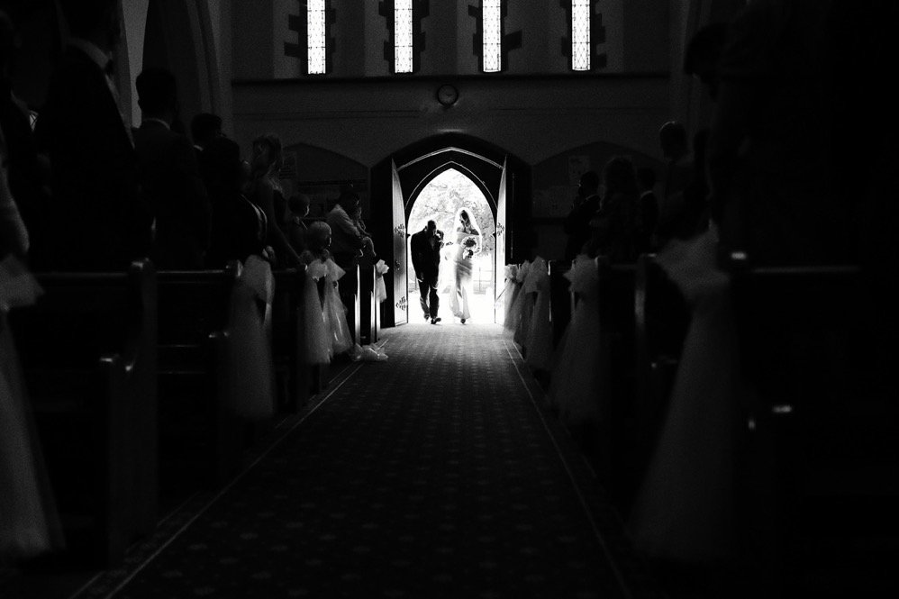 A wedding taking place inside a church, with a couple walking out of the church door while the guests stand along the pews to watch.