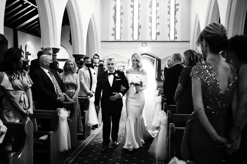 Bride and groom walking down church aisle with guests on both sides, wedding decorations, and stained glass windows