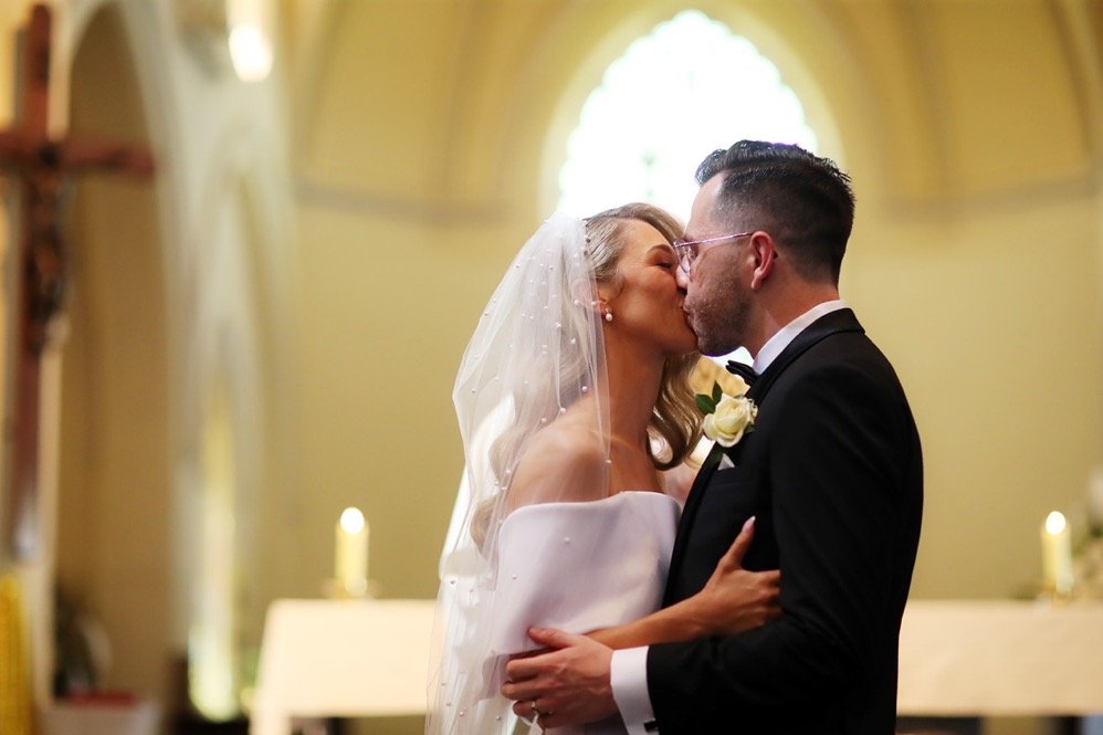 A bride and groom share a kiss during their wedding ceremony inside a church, with soft lighting and candles in the background.