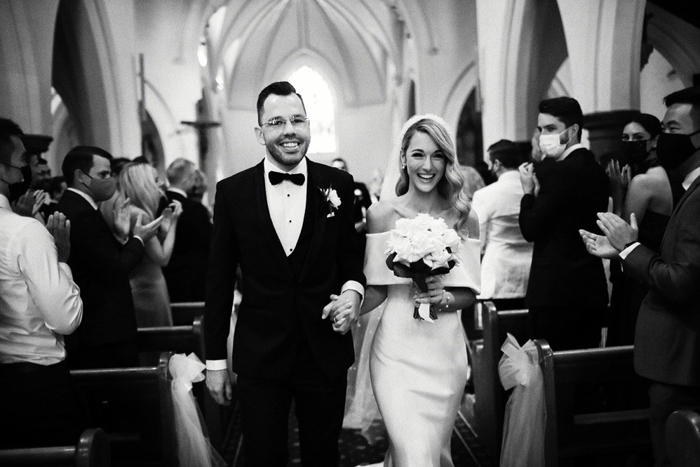 A happy bride and groom walking down the aisle in a church, surrounded by clapping guests, during their wedding ceremony. The bride wears an off-the-shoulder wedding dress and holds a bouquet of flowers, while the groom wears a tuxedo with a bow tie.