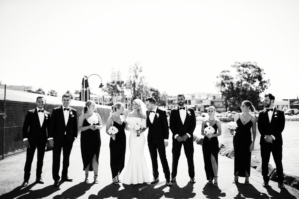 Black and white photograph of a wedding party standing outdoors on a sunny day, including a bride in a white gown surrounded by eight bridesmaids and groomsmen in formal tuxedos, all smiling.