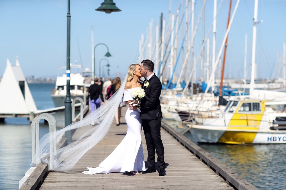 A bride and groom kissing on a dock with sailboats and a harbor in the background