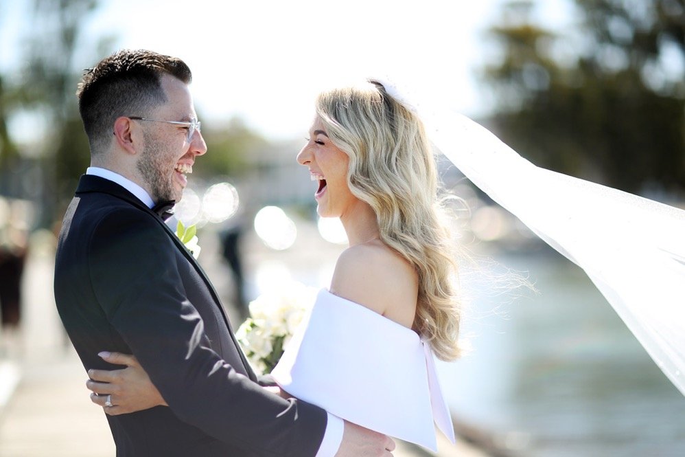 A bride and groom laughing and happiness by the water during a wedding ceremony, with the bride's veil flowing in the breeze.