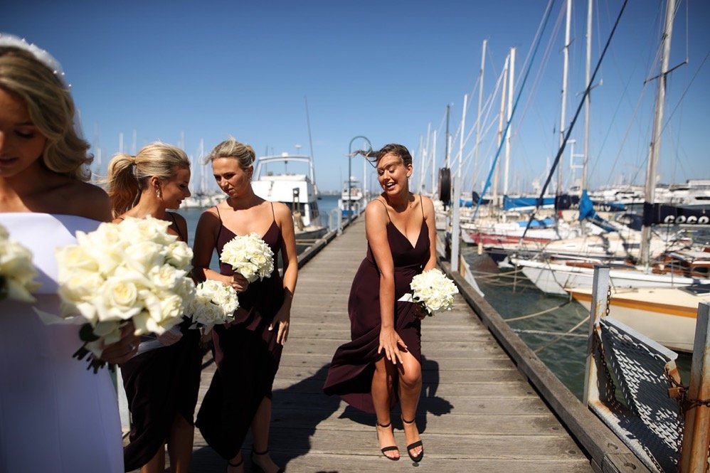 Four women standing on a marina dock, holding white bouquets, with boats and yachts in the background under a blue sky; one woman is in the foreground, lifting her leg and smiling, dressed in a dark dress.
