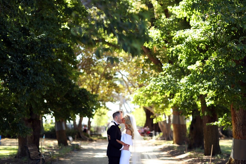 A bride and groom sharing a kiss under large leafy trees in a park.