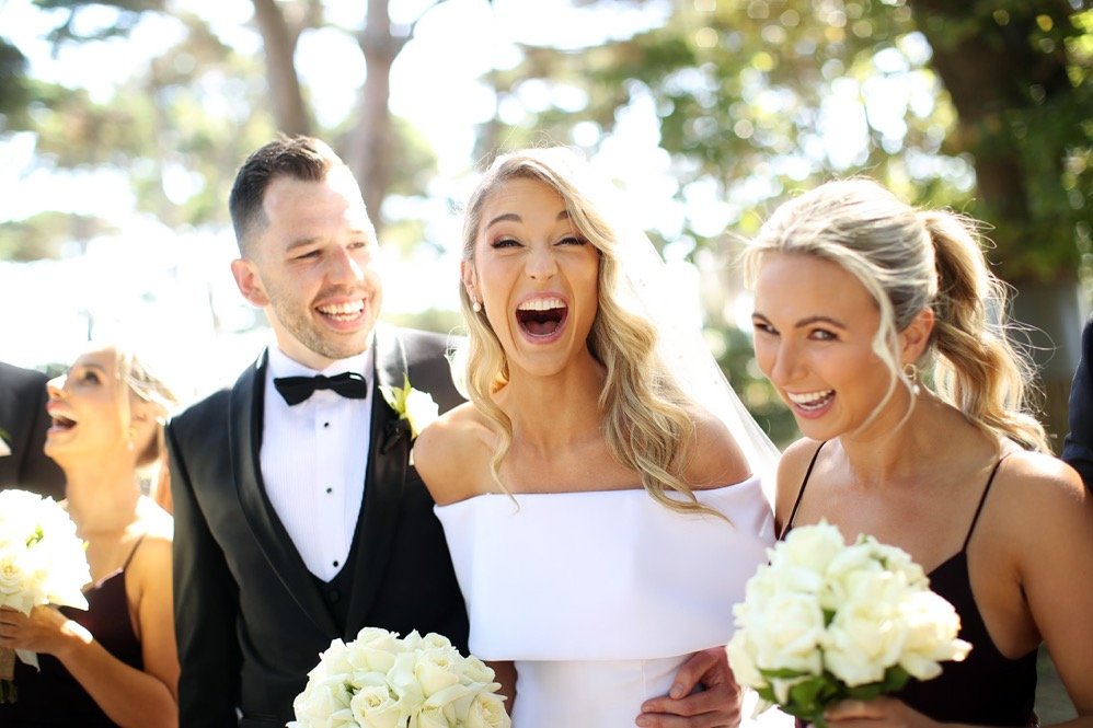 A group of people, including a bride in a white wedding dress holding a bouquet and two women with bouquets, laughing and smiling outdoors on a sunny day.