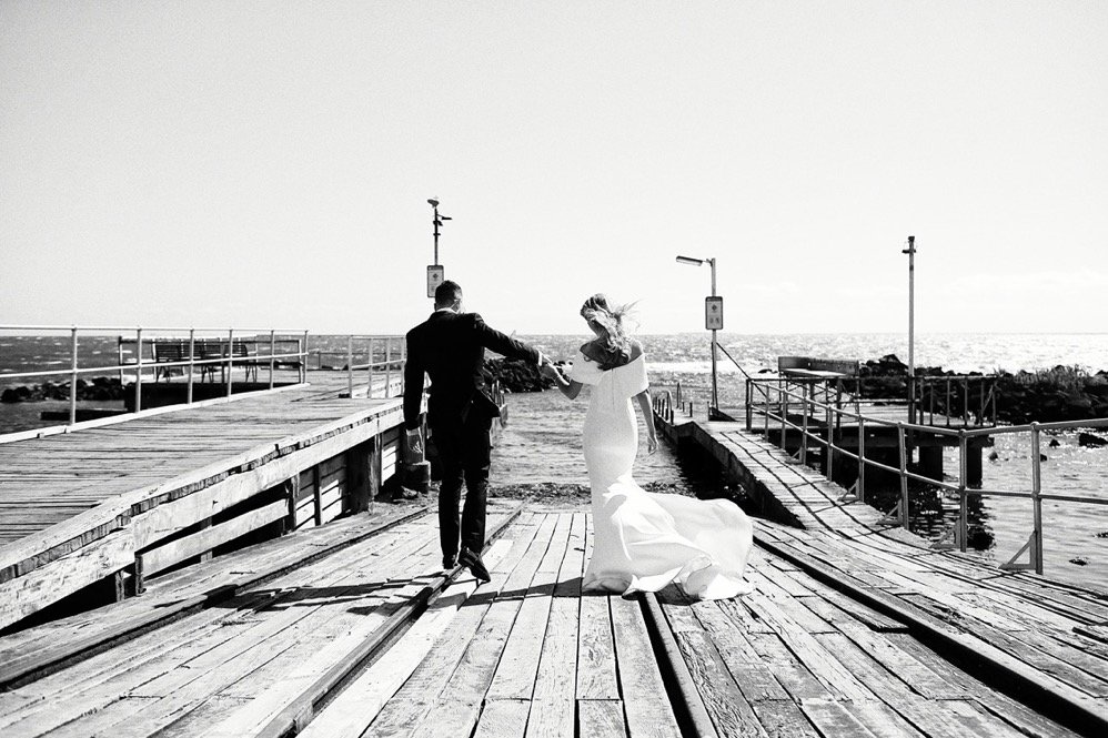 A bride and groom walking hand in hand on a wooden pier by the sea, with the bride wearing a wedding dress and the groom in a suit, the sun setting or rising in the background.
