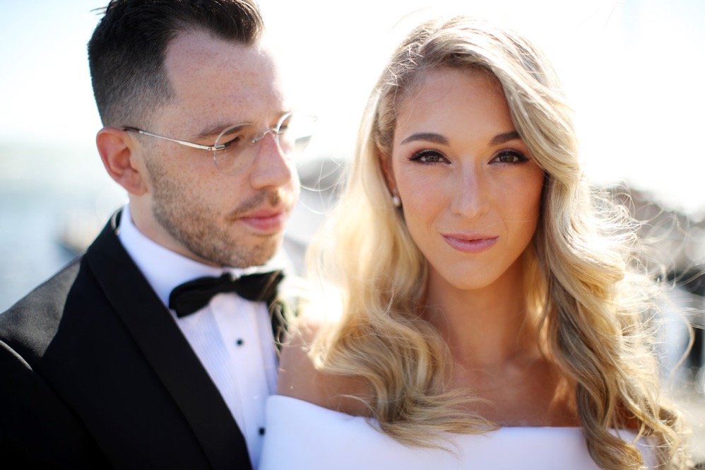 A wedding couple outdoors on a sunny day, the groom in a tuxedo and the bride in a white dress, with the bride smiling and the groom looking down.