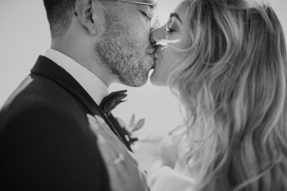 A black-and-white photo of a groom and bride kissing on their wedding day, both dressed in formal wedding attire.
