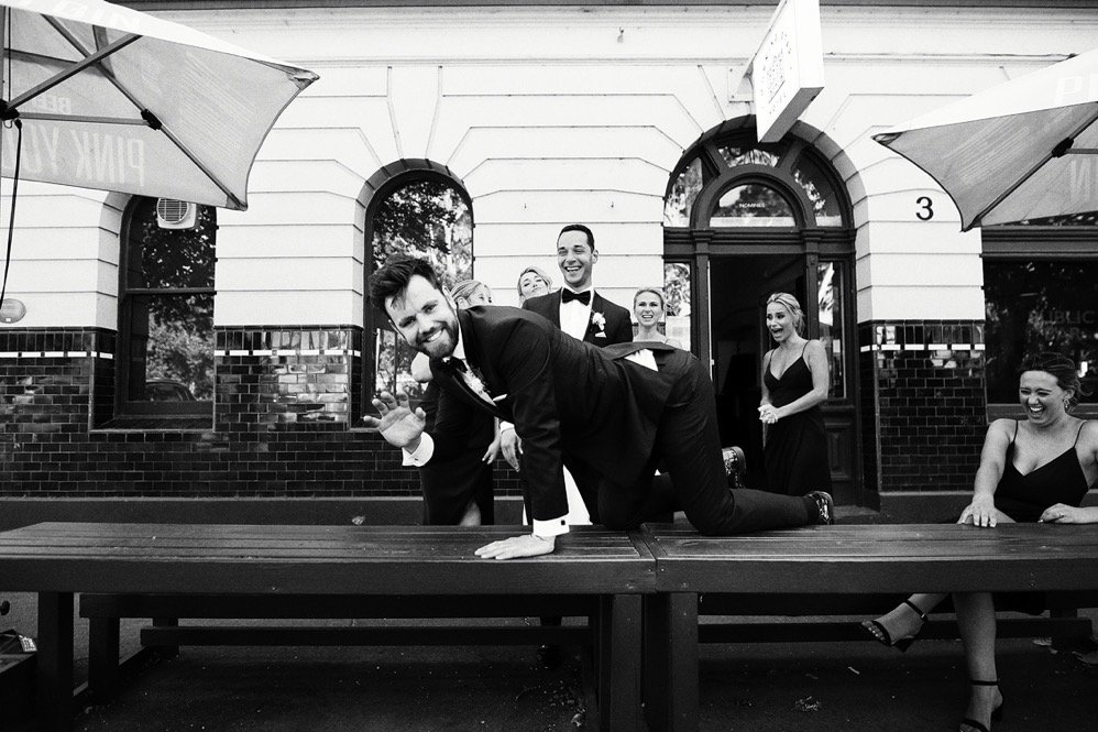 Group of people in formal attire, with one man in a suit performing a push-up on a bench outdoors, others smiling and laughing in the background.