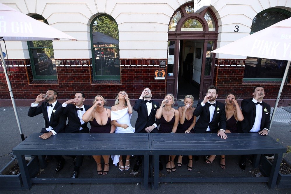 Group of nine people dressed in formal attire, drinking from glasses, standing and sitting behind a long black table outside a building with arched windows and a doorway.