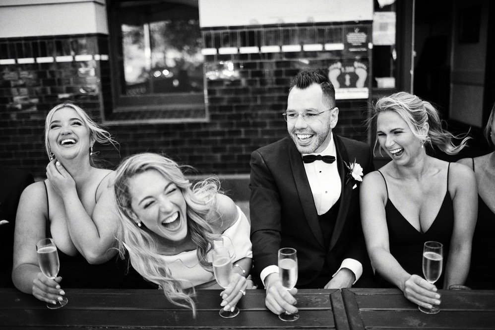 Group of people, four women and one man, laughing and celebrating at a social gathering, with drinks in hand, in a venue with dark brick walls.
