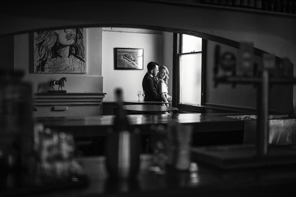 A woman and man are sitting close together by a window, sharing an intimate moment, with a glass of wine nearby in a cozy, art-decorated room.