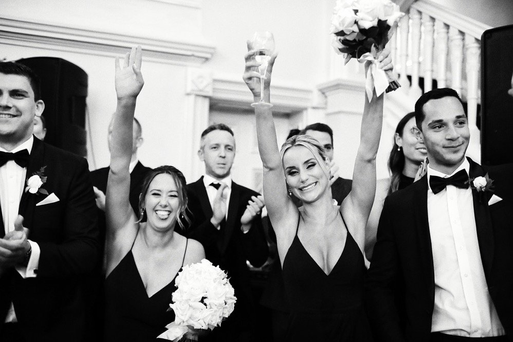 Woman in black dress holding bouquet and raising glass at wedding celebration, surrounded by smiling guests in formal attire.