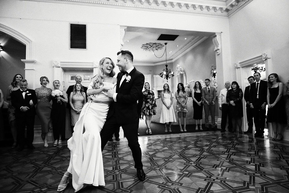 Black and white photo of a bride and groom dancing at their wedding reception, surrounded by guests in an elegant hall.