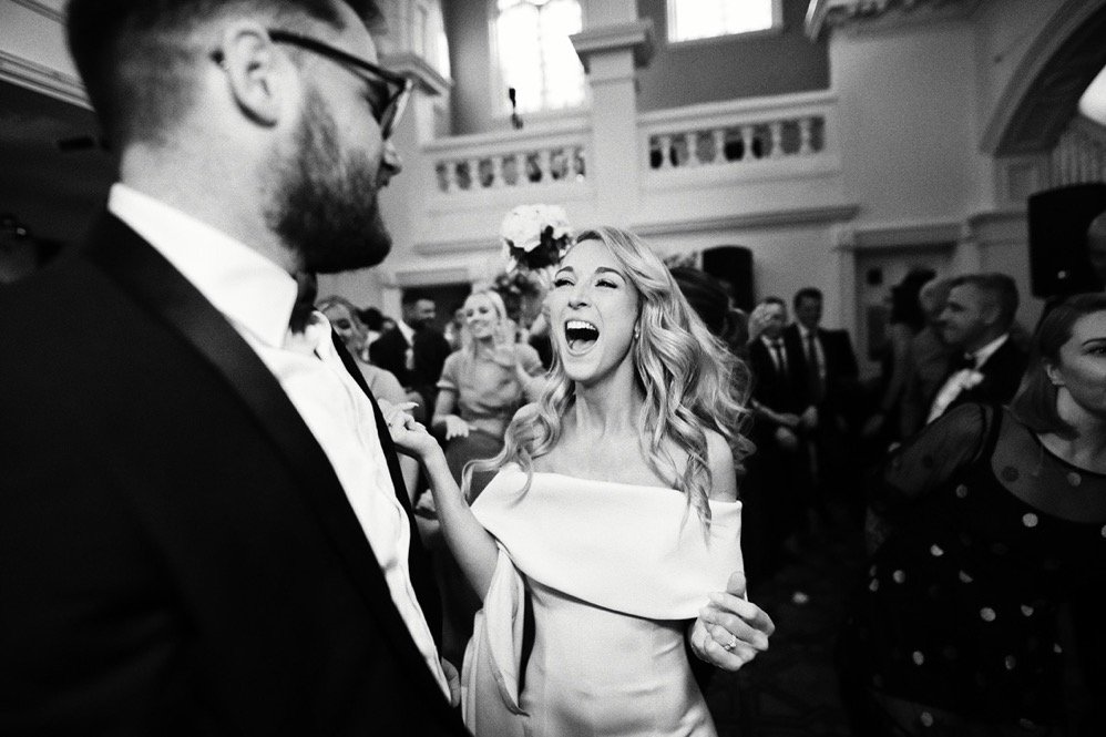 A woman in an off-shoulder wedding dress laughing joyfully at a wedding reception, surrounded by guests in a decorated hall.