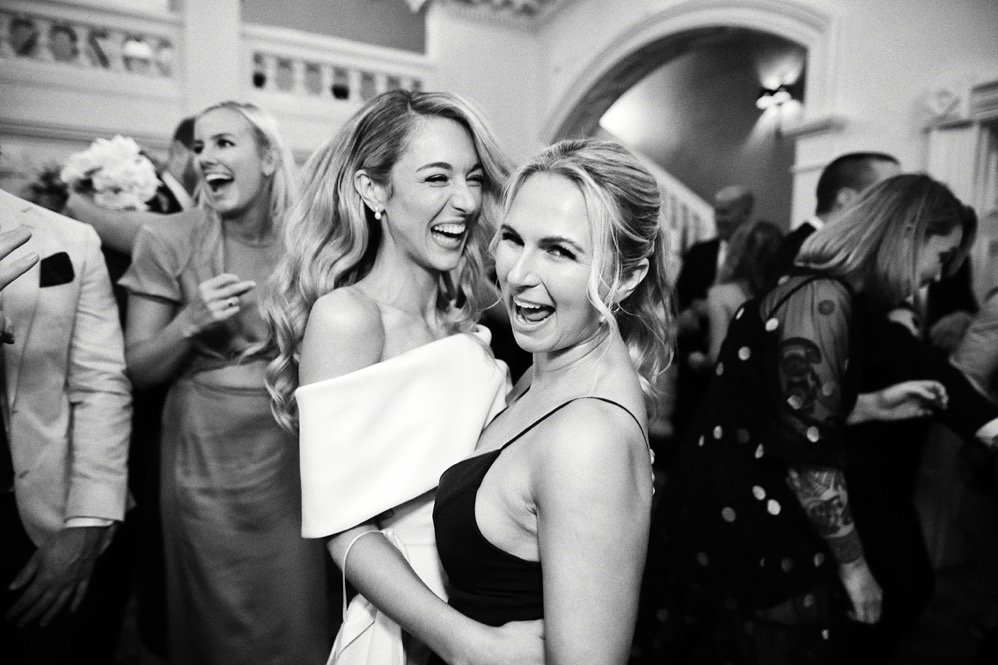 Two women in formal attire smiling and laughing together at a party or wedding reception.